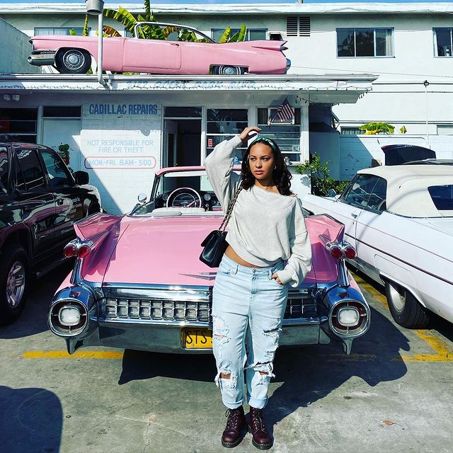 Jasmine Cephas Jones in a white t-shirt and light blue jeans and black boots posing in front of her pink car.